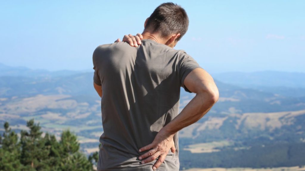 man standing outside on a mountain grabbing is neck and low back in pain