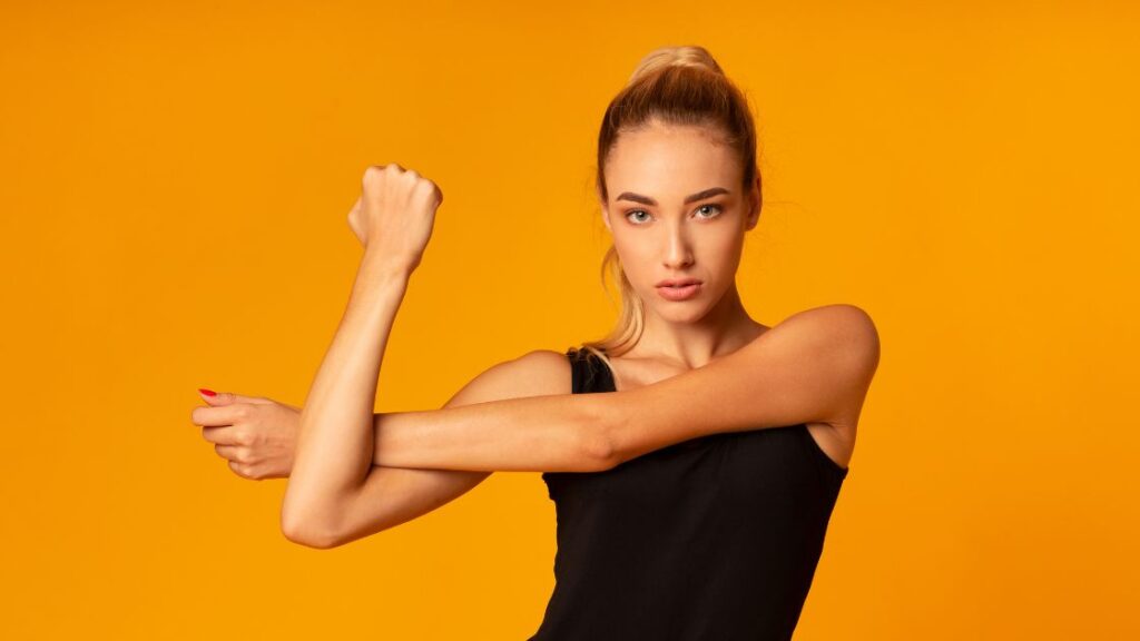 woman in a black shirt stretching arm across her body in front of a yellow background