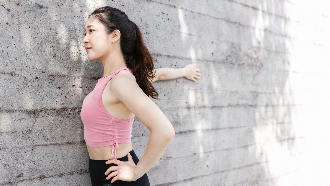 woman in a pink top stretching one arm on a grey wall