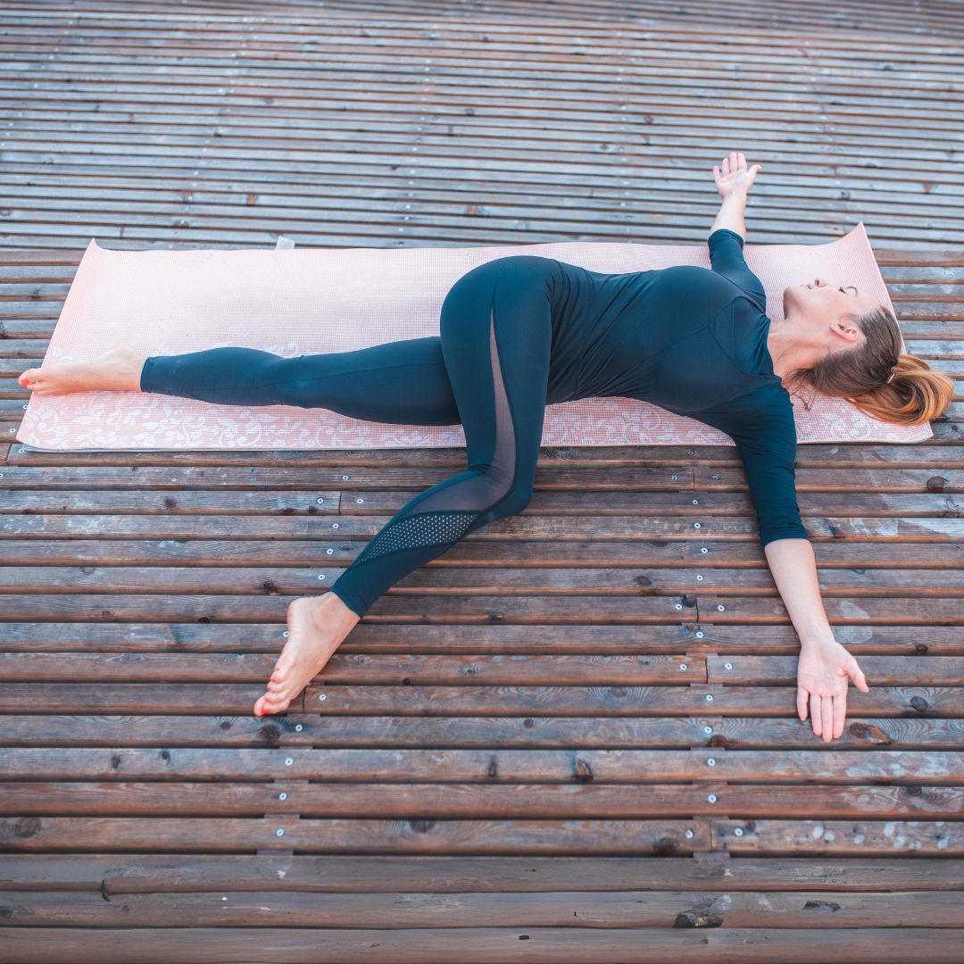 woman doing supine spinal twist wearing all black on a pink yoga mat on a wood deck