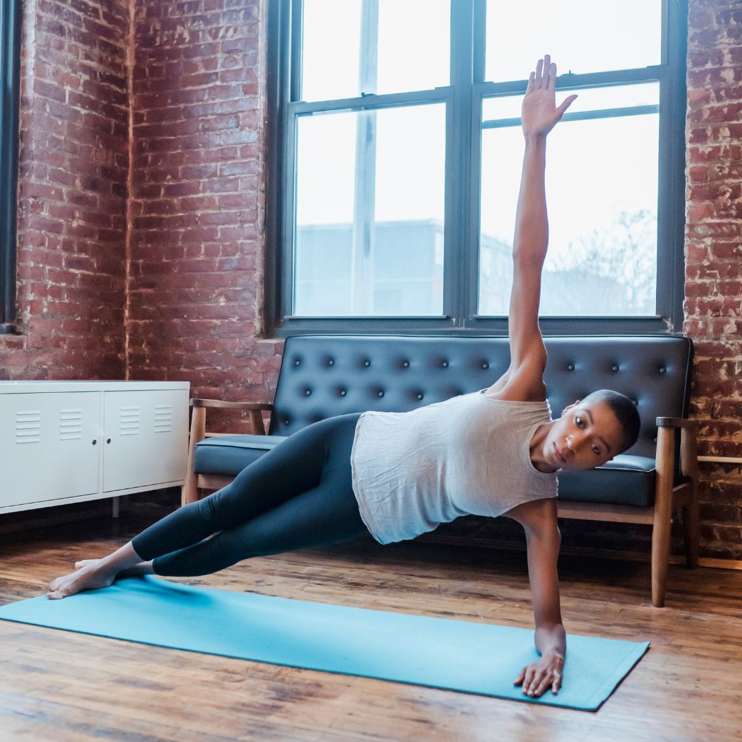 woman in a grey tank doing a T-Plan on a blue yoga mat in front of a brick wall and black couch