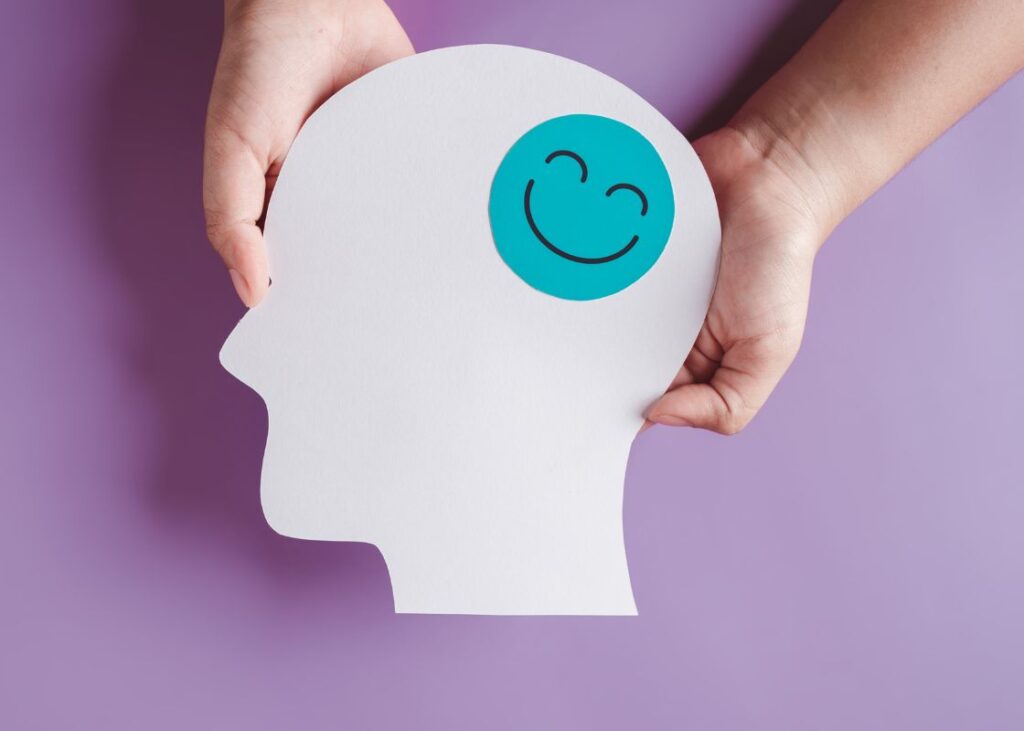 hands holding a cutout of a head with a smiley face on the brain